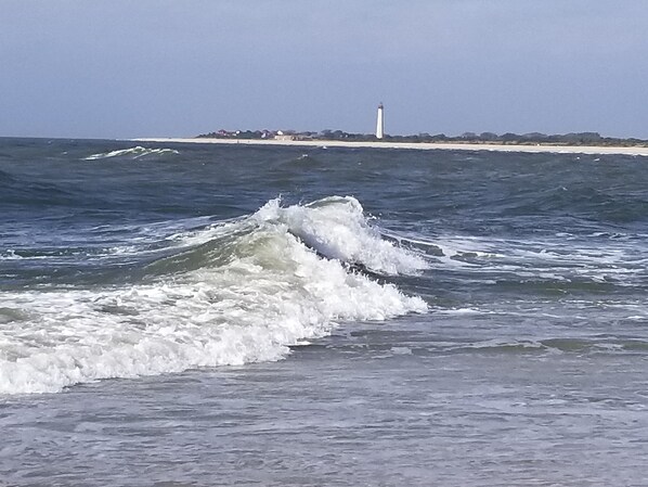 A coastal scene with waves approaching the shore, a lighthouse in the distance, and a sandy beach with sparse vegetation. - Conveniently located Wildwood Crest Condo!  Enjoy the Jersey Shore! (Wildwood Crest)