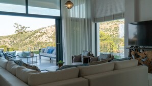 A modern living room with a large sectional sofa, a flat-screen TV, and a view of a hilly landscape through large windows. - Eliandreia Luxury Villa (Lefkada)