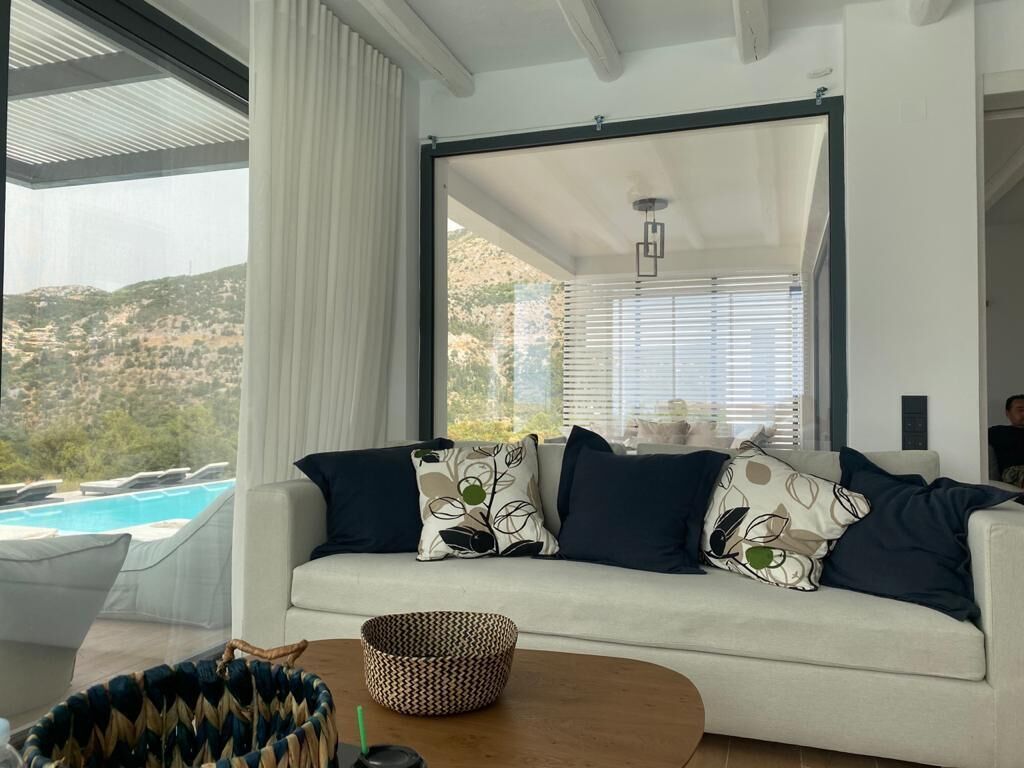 Un salon moderne avec un canapé blanc, des coussins à motifs, un panier en osier et une vue sur une piscine et des collines.