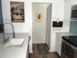 A modern kitchen with white cabinetry, a stainless steel sink, a black stove, and a microwave. There is a door with a colorful artwork and a small wine rack with glasses. - Sea Colony-Perfect Beach Getaway-Newly Renovated (Bethany Beach)