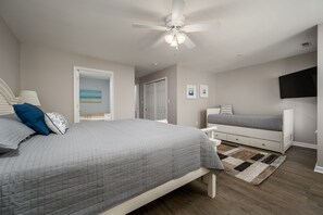 Quarto com cama, mesas de cabeceira, ventilador no teto, televisão e porta para outro cômodo.