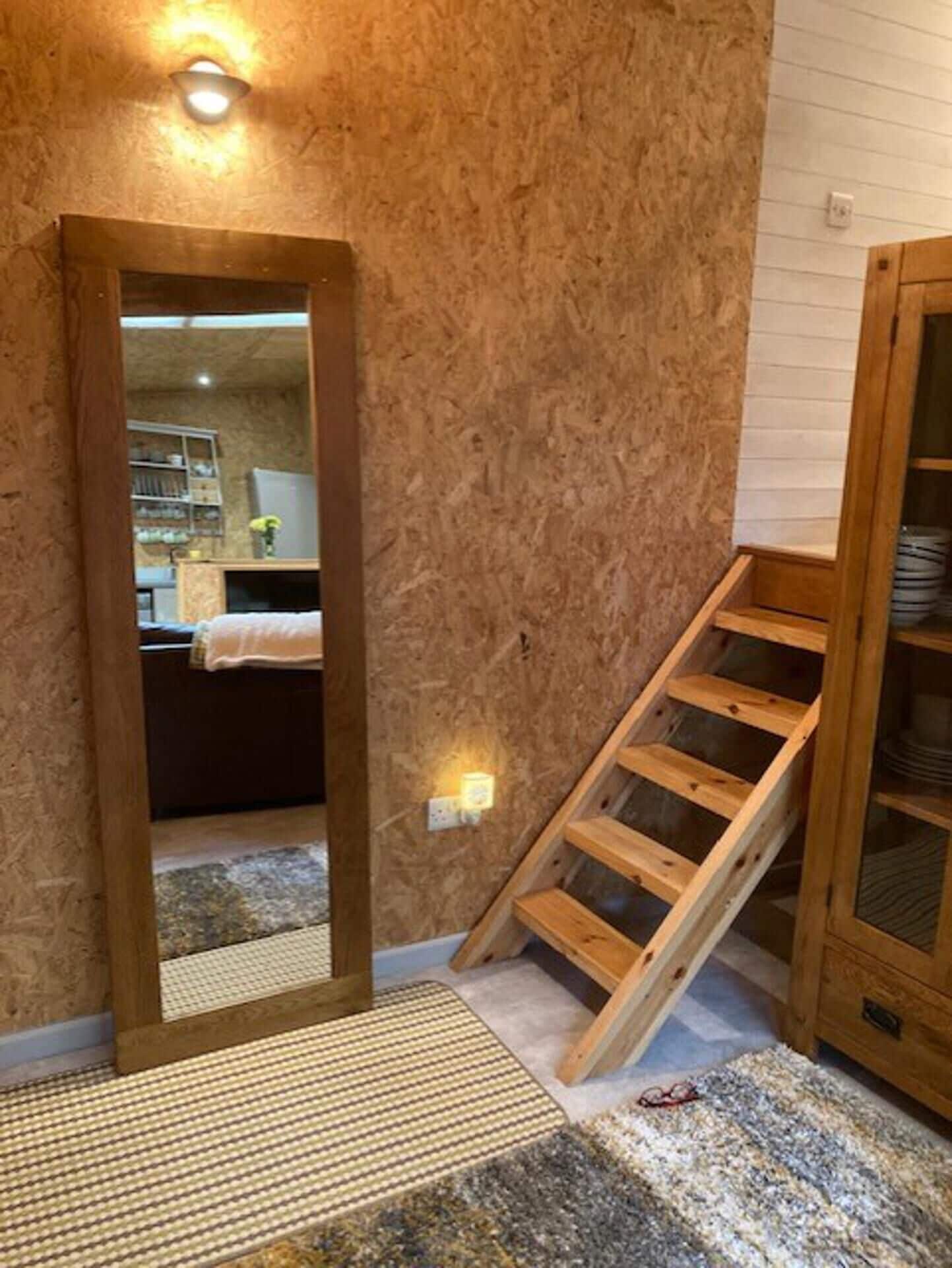 A room with a wooden staircase, a mirror, and a view into another room with a bed and shelves.