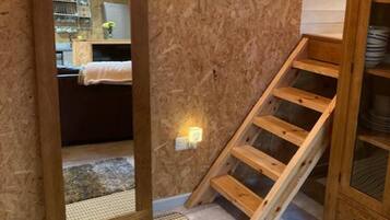 A room with a wooden staircase, a mirror, and a view into another room with a bed and shelves.
