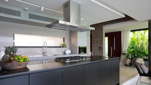 A modern kitchen with a central island, stainless steel appliances, and a large window providing natural light. - Muscat 3-Bedroom Villa with Beach Access (Koh Samui)