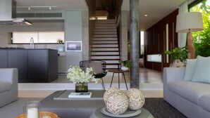 A modern living room with a sofa, a coffee table with decorative items, a staircase, and a kitchen area in the background. - Gamay 3-Bedroom Villa with Beach Access (Koh Samui)