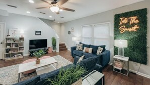 Uma sala de estar moderna com um ventilador de teto, uma televisão, um conjunto de sofás, uma mesa de centro e uma placa na parede com a inscrição "Into The Sunset".