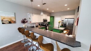Cozinha moderna com ilha central, banquetas, armários verdes e eletrodomésticos de aço inoxidável.