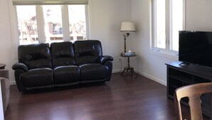 A living room with a black leather sofa, a wooden chair, a TV on a stand, a lamp, and large windows with a view of buildings outside. - Lake Mille Lacs Cabin. Onamia (Onamia)