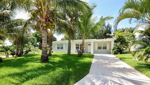 A single-story house with a white exterior, surrounded by lush green grass and palm trees. - Driftwood Way~ No Checkout Chores (Jensen Beach)