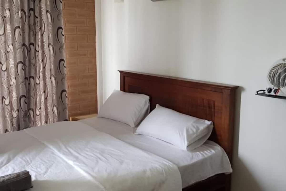 A neatly made bed with white linens and two pillows, a wooden headboard, a wall-mounted fan, and a curtain with a wavy pattern.