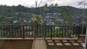 A balcony with a metal railing, a tiled floor, and a view of a rural landscape with houses and greenery. - Hotel Coldworld (Nuwara Eliya)