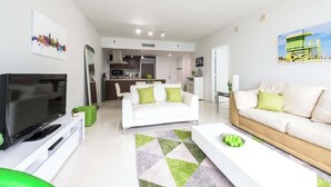 A modern living room with a flat-screen TV, a white sofa, and a dining area with a kitchen in the background.