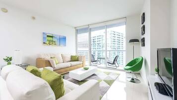 Una sala moderna con un sofá, una mesa de centro y un televisor. Hay un balcón con vistas a edificios.