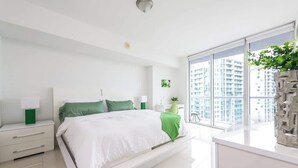 Un dormitorio moderno con cama, mesitas de noche, escritorio y un ventanal con vistas a rascacielos.