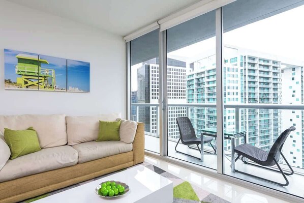 Un sal ón moderno con un sofá color beige, cojines verdes, una mesa de centro blanca con un cuenco de manzanas y un ventanal con vistas a la ciudad.