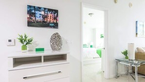 Una habitación de hotel moderna con una pared blanca, un televisor de pantalla plana montado sobre un mueble blanco, una mesa de centro de vidrio y una cama con ropa de cama verde.
