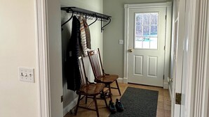 Un couloir avec un porte-manteau, deux chaises en bois, une porte avec une fenêtre et un sol carrelé.