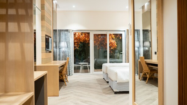 Una habitación de hotel moderna con una cama grande, un escritorio y una silla. Hay una puerta corrediza de vidrio que da a un balcón con hojas otoñales.
