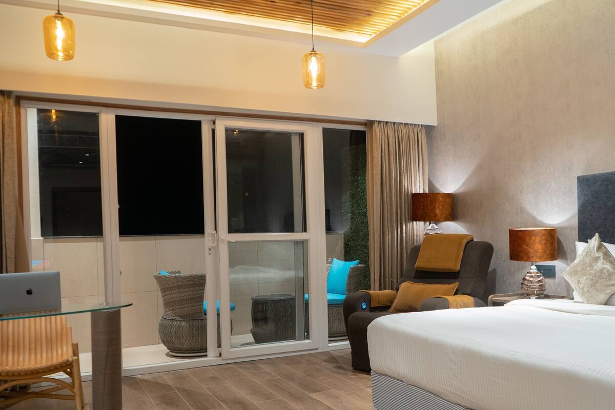 A hotel room with a bed, two chairs, a desk with a laptop, and a sliding glass door leading to a balcony.