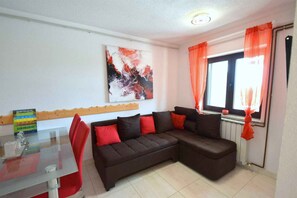 Un salon avec une table en verre, un canapĂ© marron agrĂ©mentĂ© de coussins rouges, une fenĂȘtre avec des rideaux orange et un tableau abstrait accrochĂ© au mur.