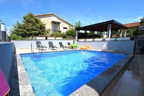 A swimming pool with lounge chairs, a covered seating area, and a building in the background.