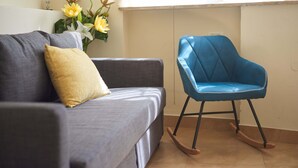 Una sala de estar con un sofá gris, un cojín amarillo, un sillón mecedor azul y un jarrón con flores amarillas.