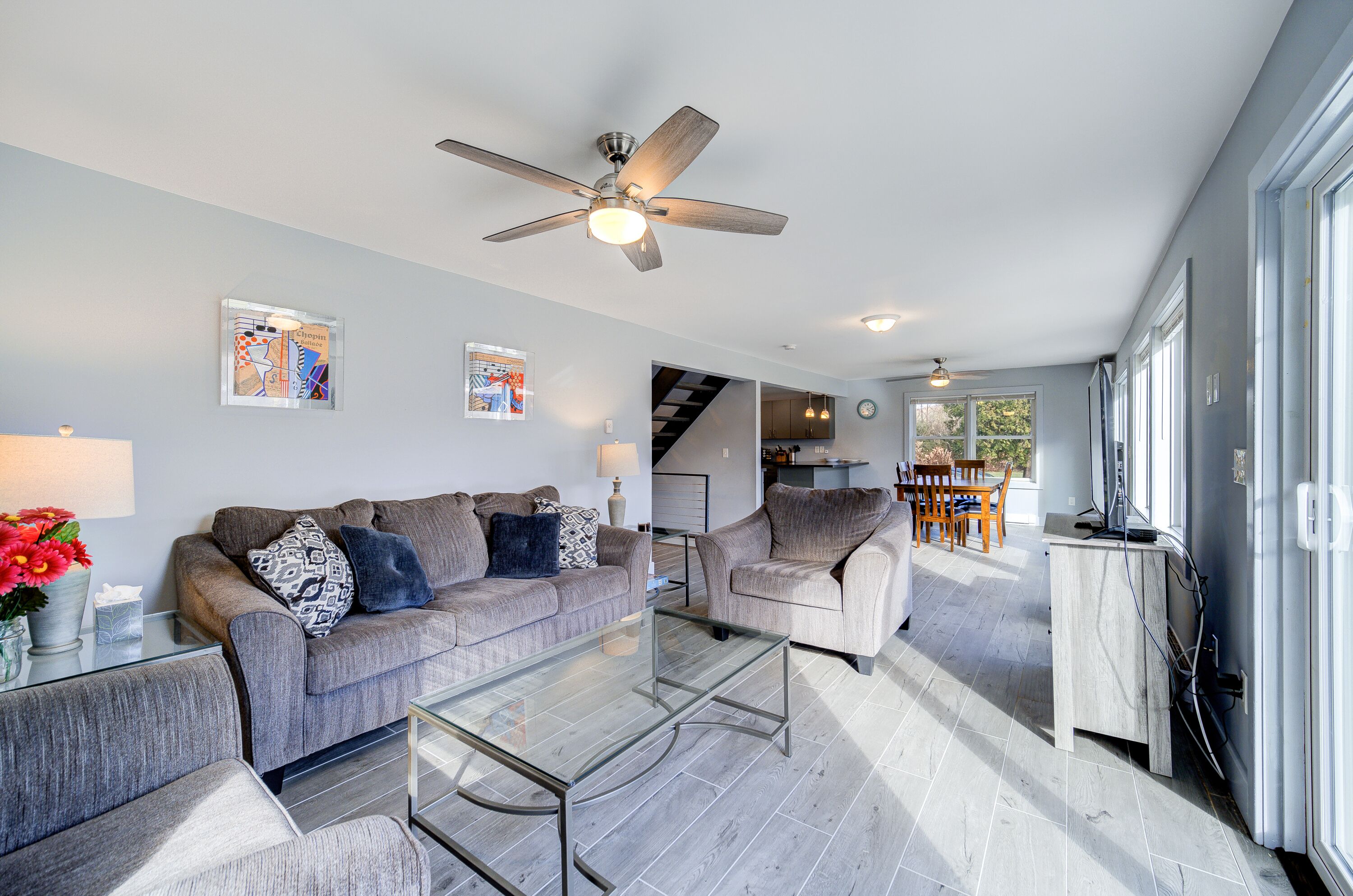 A modern living room with a grey sofa set, a glass coffee table, and a ceiling fan. There is a staircase leading to an upper level, and a dining area with a table and chairs.