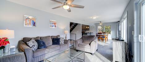 A modern living room with a grey sofa set, a glass coffee table, and a ceiling fan. There is a staircase leading to an upper level, and a dining area with a table and chairs.