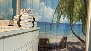 A bathroom with a beach-themed shower curtain, a stack of towels, and a beach scene mat.