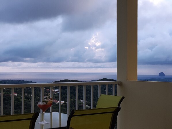 A balcony with a table and chairs, overlooking a coastal landscape with a cloudy sky.