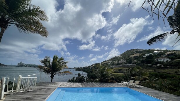 Una terraza de madera con piscina, palmeras y vistas a una zona costera con casas.
