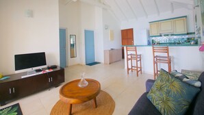 Amplia sala de estar con televisor, una mesa de centro de madera y un sofá con un cojín decorativo. Se ve un área de cocina al fondo con armarios de madera y una encimera.