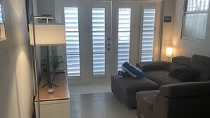 Un salon avec un lampadaire, un canapé et une table basse. Il y a deux portes équipées de stores, un ventilateur de plafond et un appareil de climatisation.