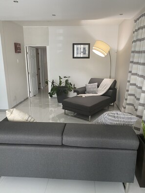 A modern living room with a grey sofa, a black armchair, and a floor lamp. There is a potted plant and a framed picture on the wall.