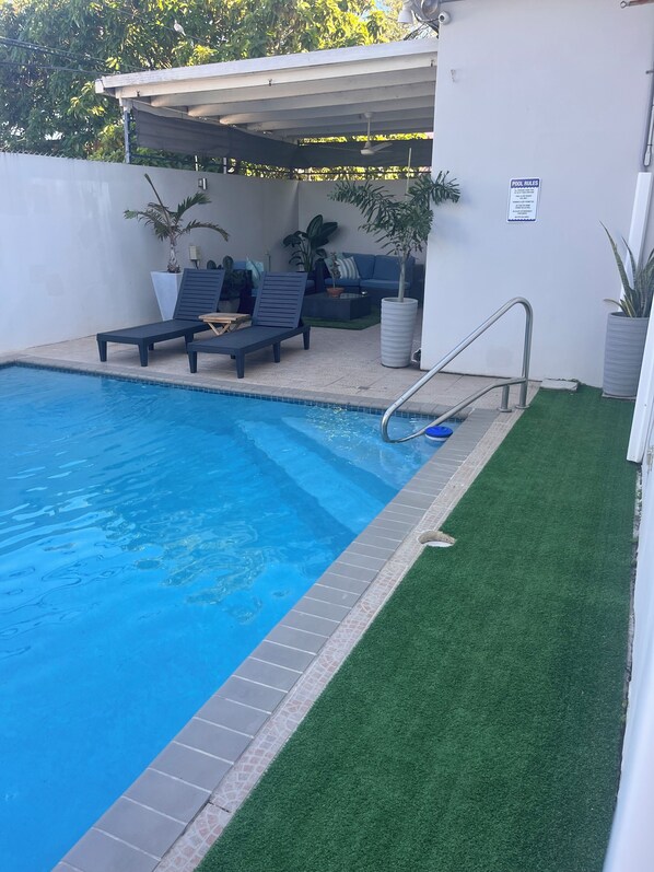 Una piscina con una escalera metálica, rodeada de césped artificial y sillas de descanso.