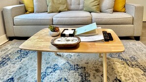 A living room with a grey sofa, yellow and grey cushions, a wooden coffee table with a bowl, remote controls, and a macrame wall hanging. - Welcome home! Your beach cottage awaits! (Fort Lauderdale)