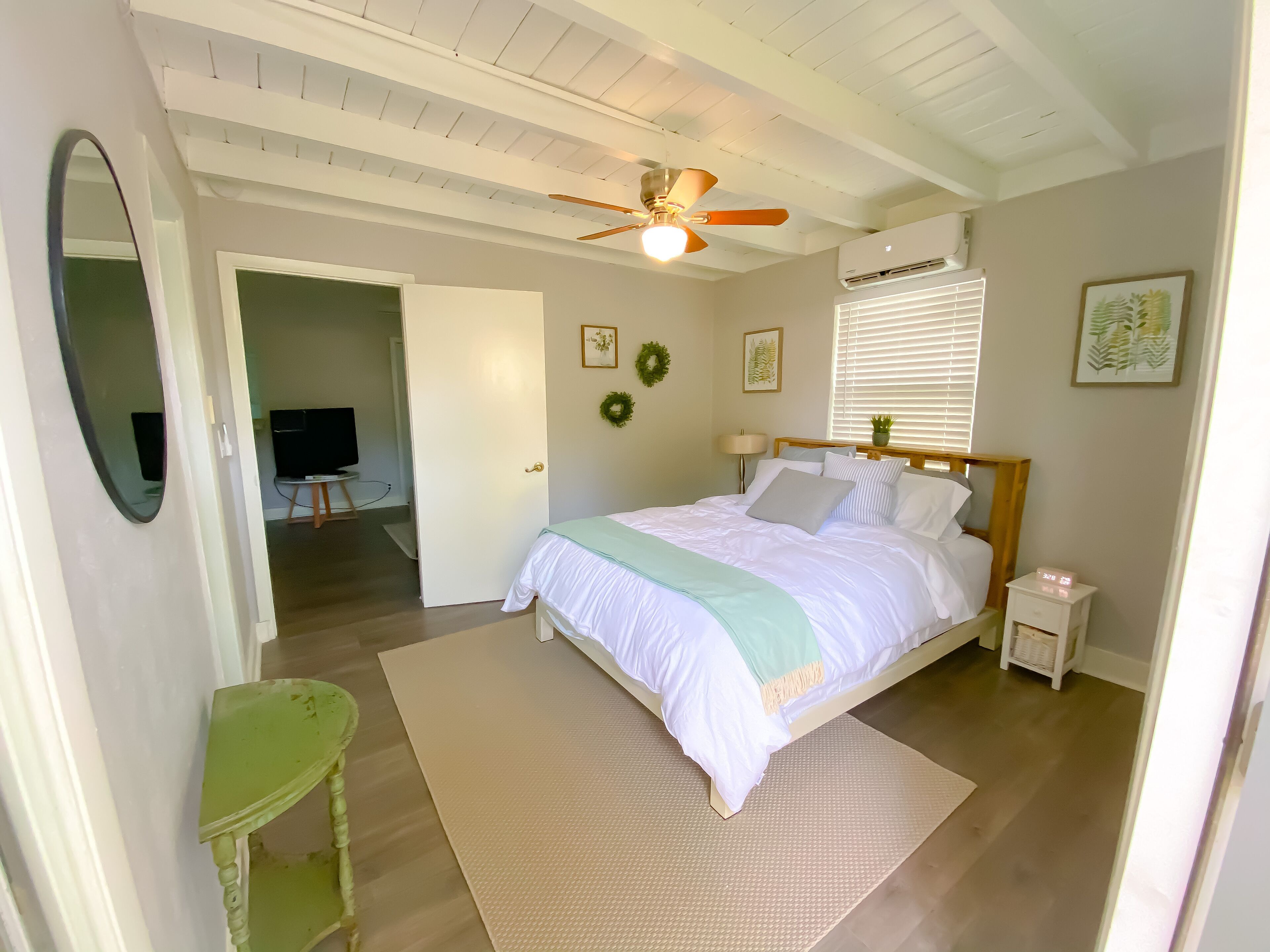 Ein Schlafzimmer mit einem Bett, einem Deckenventilator, einem Nachttisch und einem Fernseher im angrenzenden Raum. Eine Tür führt in einen weiteren Raum.