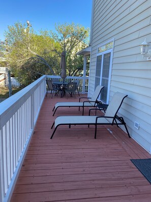 A wooden deck with two lounge chairs and a table, a white railing, and a house with a window and a light fixture. - Cozy home within walking distance to the beach. Golf cart available for x-fee (Surfside Beach)