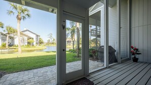 Eine Holzdielenfläche mit Fußmatte, einer Tür zu einer überdachten Terrasse und Blick auf einen angelegten Garten mit Palmen und einem Teich.