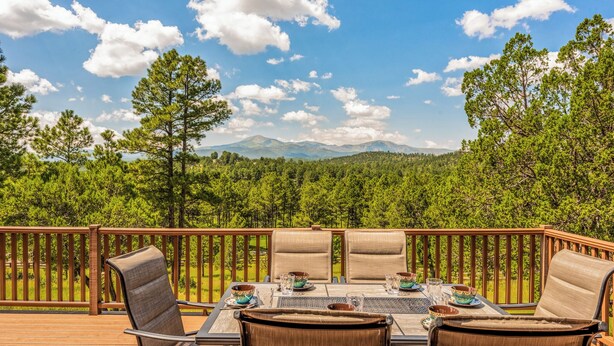 A deck with a table set for four, surrounded by chairs, overlooking a forest and mountains.