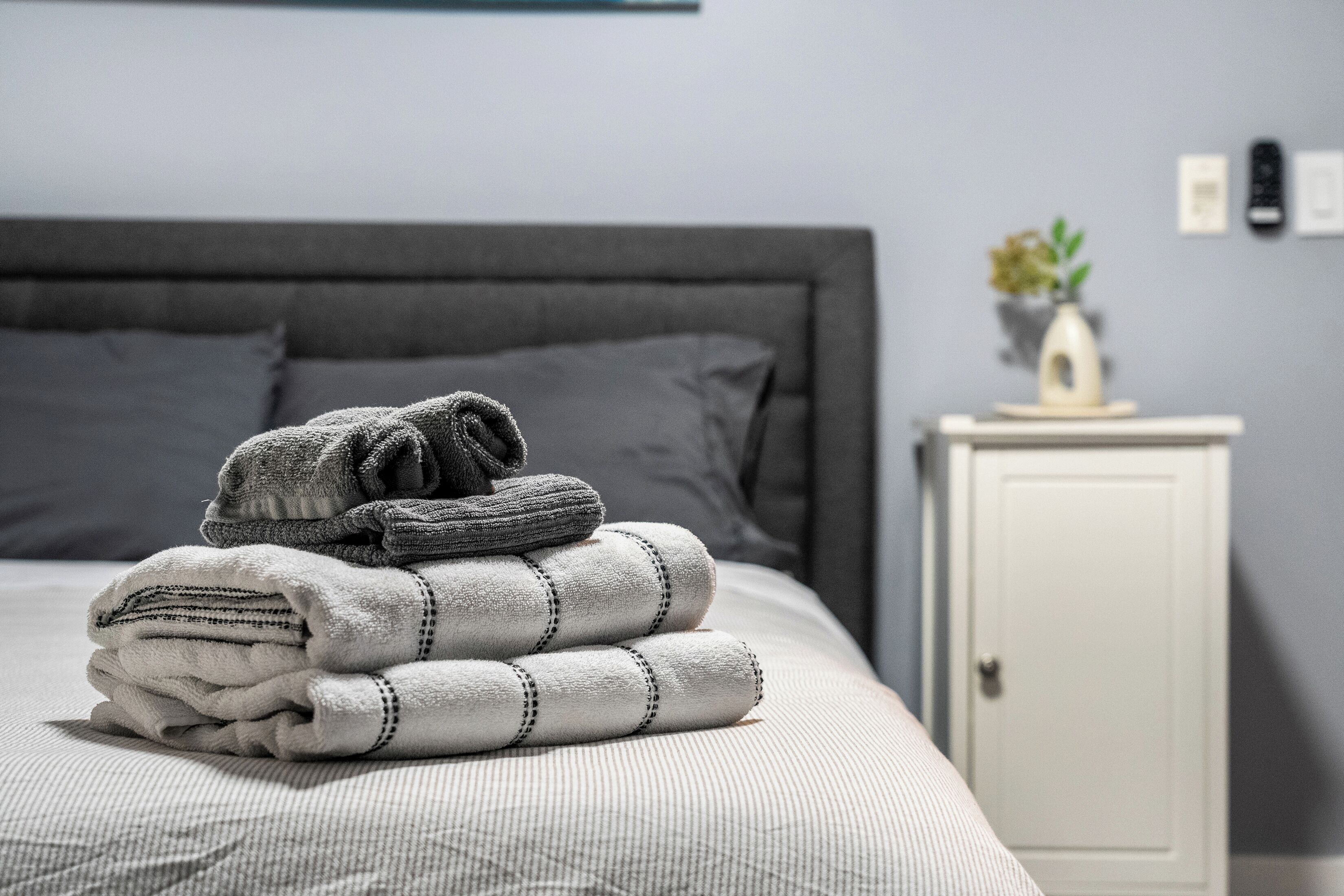 A neatly made bed with folded towels and a bedside table with a vase and a plant.