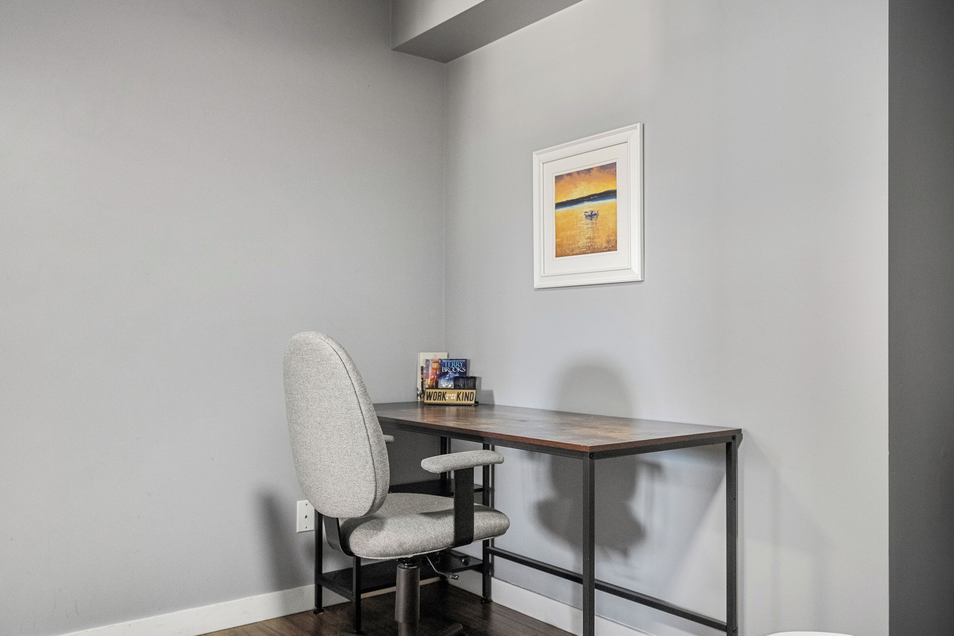 Un bureau d’angle avec une chaise de bureau grise, un bureau en bois et un tableau encadré accroché au mur.