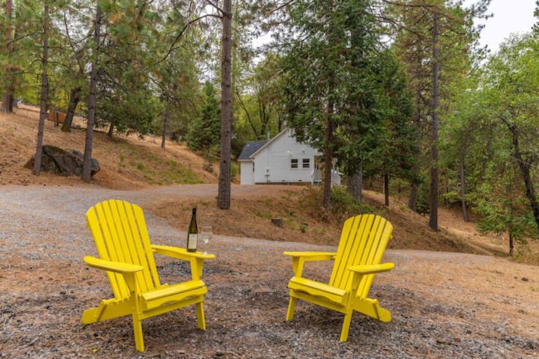 Twee gele Adirondack stoelen staan op een grindpad, met op de achtergrond een huis omringd door bomen.