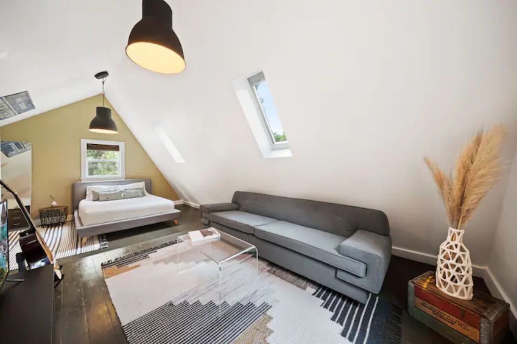 A modern loft bedroom with a sloped ceiling, a gray sofa, a bed with a white headboard, a glass coffee table, and a small window with a view.