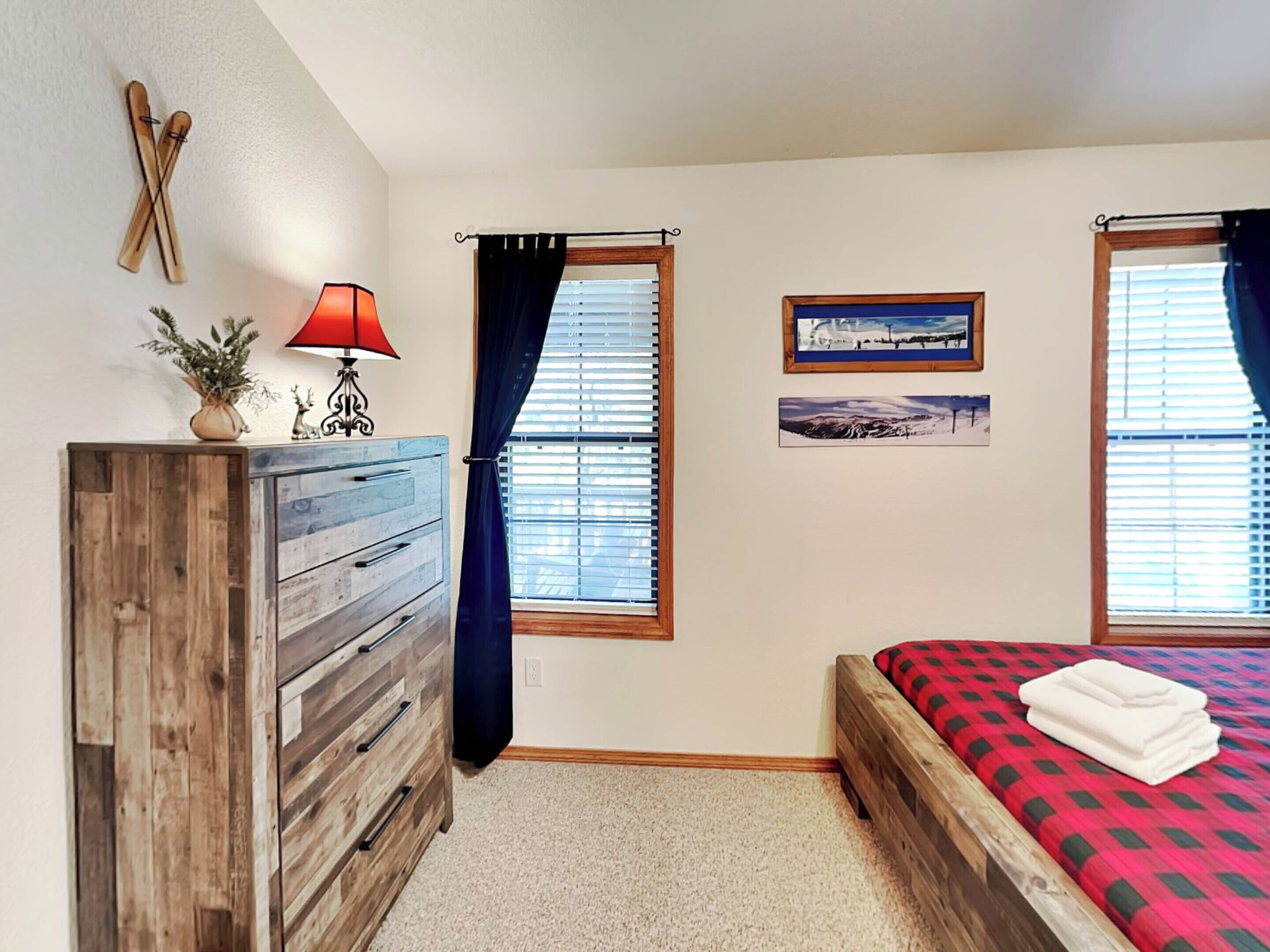 A bedroom with a wooden dresser, a bed with a red and black checkered bedspread, two windows with blinds, a wall-mounted lamp, and a framed picture.