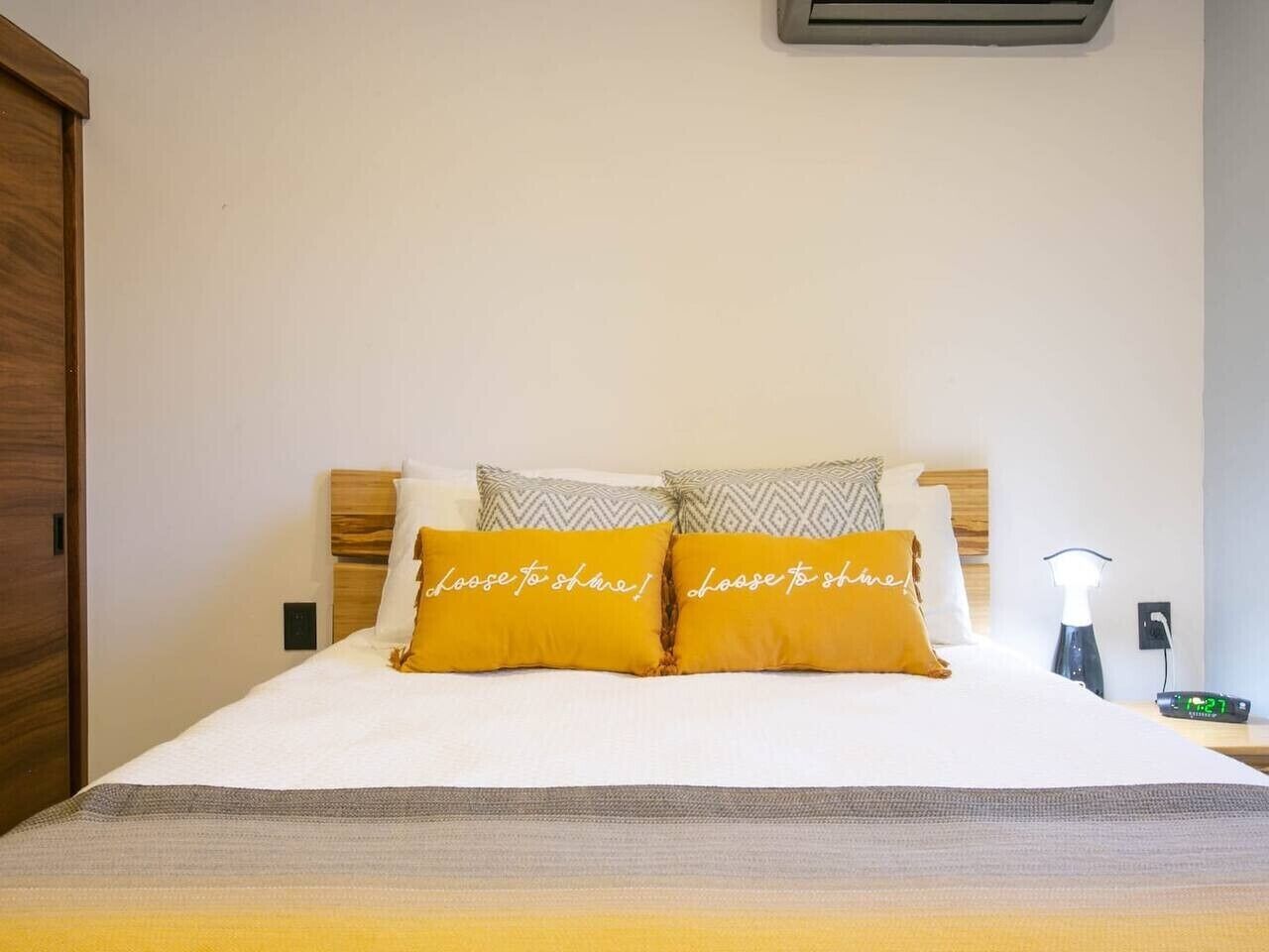 A neatly made bed with two pillows, a bedside table with a lamp, and a wall-mounted air conditioner.