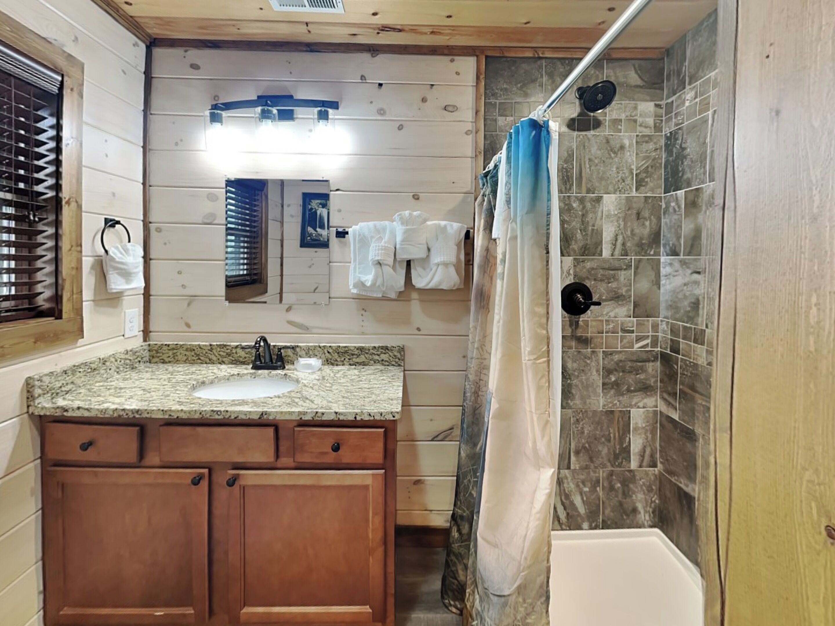 A bathroom with a granite countertop sink, wooden cabinets, a mirror, a shower with a curtain, and tiled walls.