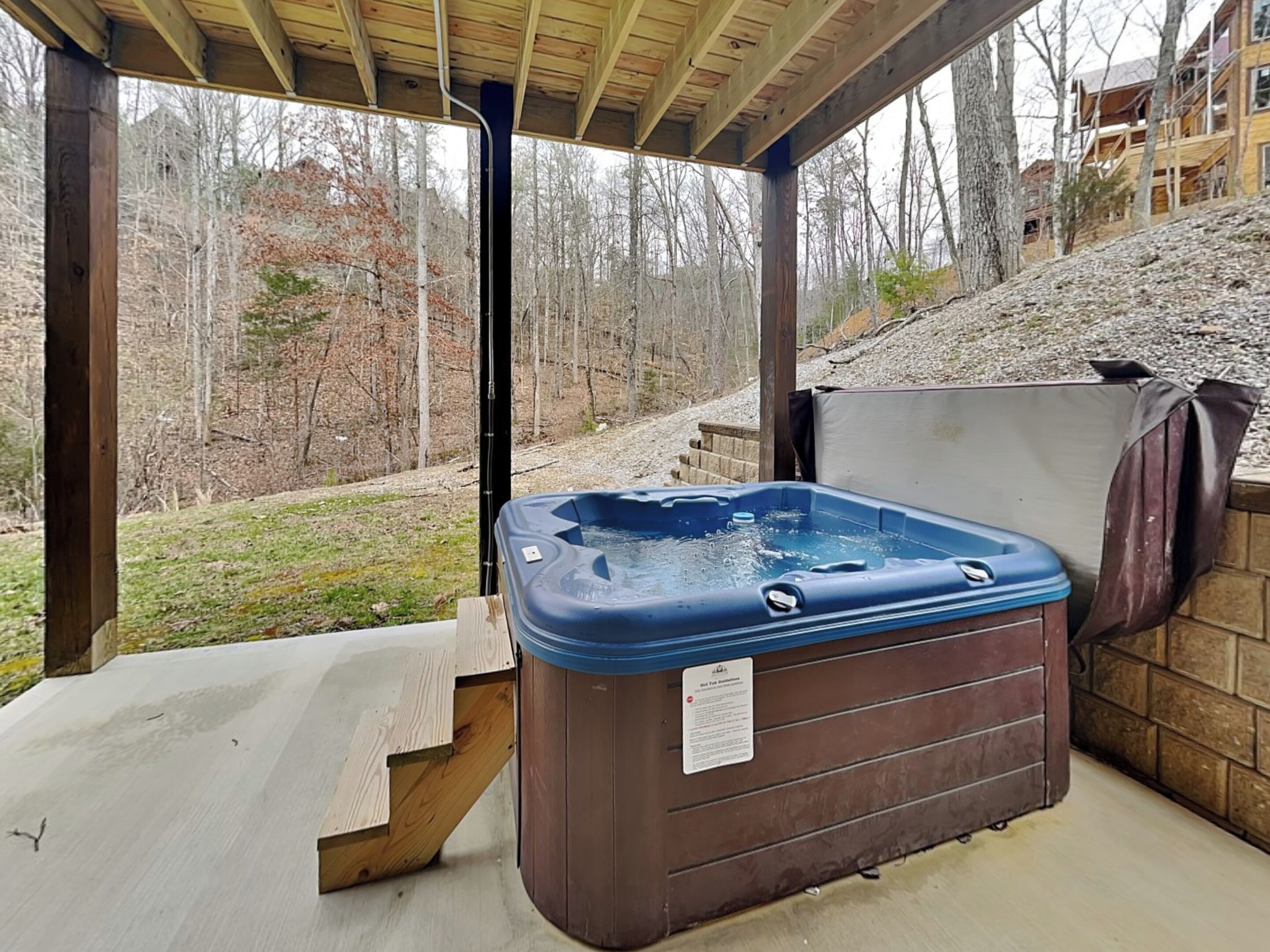 Un bain à remous situé dans un espace extérieur abrité, avec vue sur une zone boisée et un bâtiment en arrière-plan.