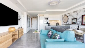 A modern living room with a sofa, a TV, and a kitchen in the background. - Apartment Royal Sunset Loggia (Palm-Mar)
