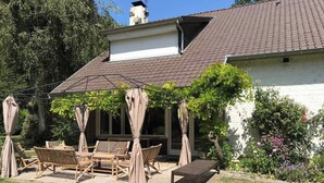 A patio with wooden furniture and a pergola with curtains, surrounded by a garden and a white building with a tiled roof. - Country house nestled in a green setting 15 minutes from Lille (Sainghin-en-Mélantois)